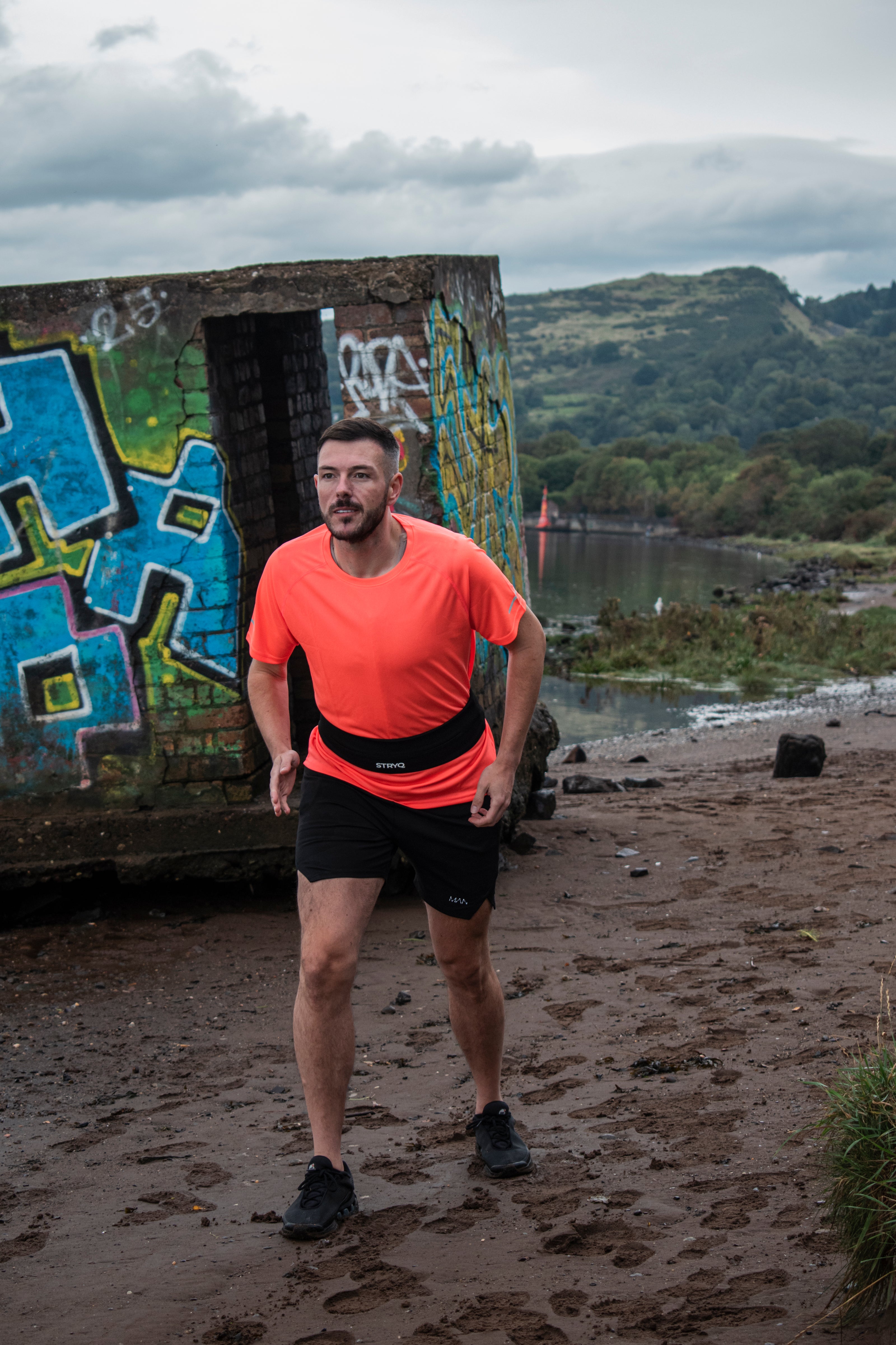 Man in athletic wear standing on a beach with graffiti and water in the background wearing a running vest